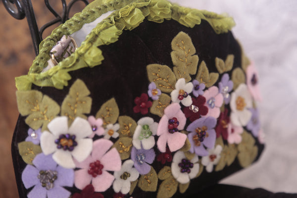 A stylish velvet purse decorated with colorful felt flowers and beads, perfect for adding a pop of charm to any outfit.
