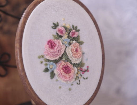 Embroidery art displaying three pastel peach roses elegantly stitched on a circular hoop canvas along with Tiny Blue Flowers and Rose buds.